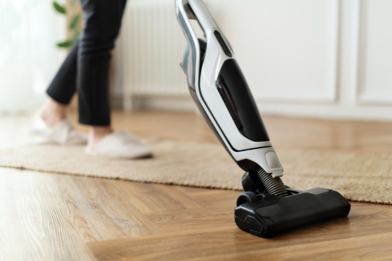 Person vacuuming wooden floor with a white cordless stick vacuum beside a woven rug in a home cleaning scene