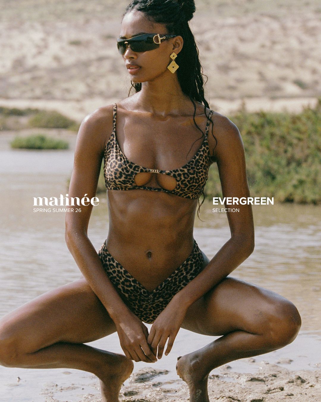 Woman model in leopard-print bikini and sunglasses squatting on a sandy beach by shallow water