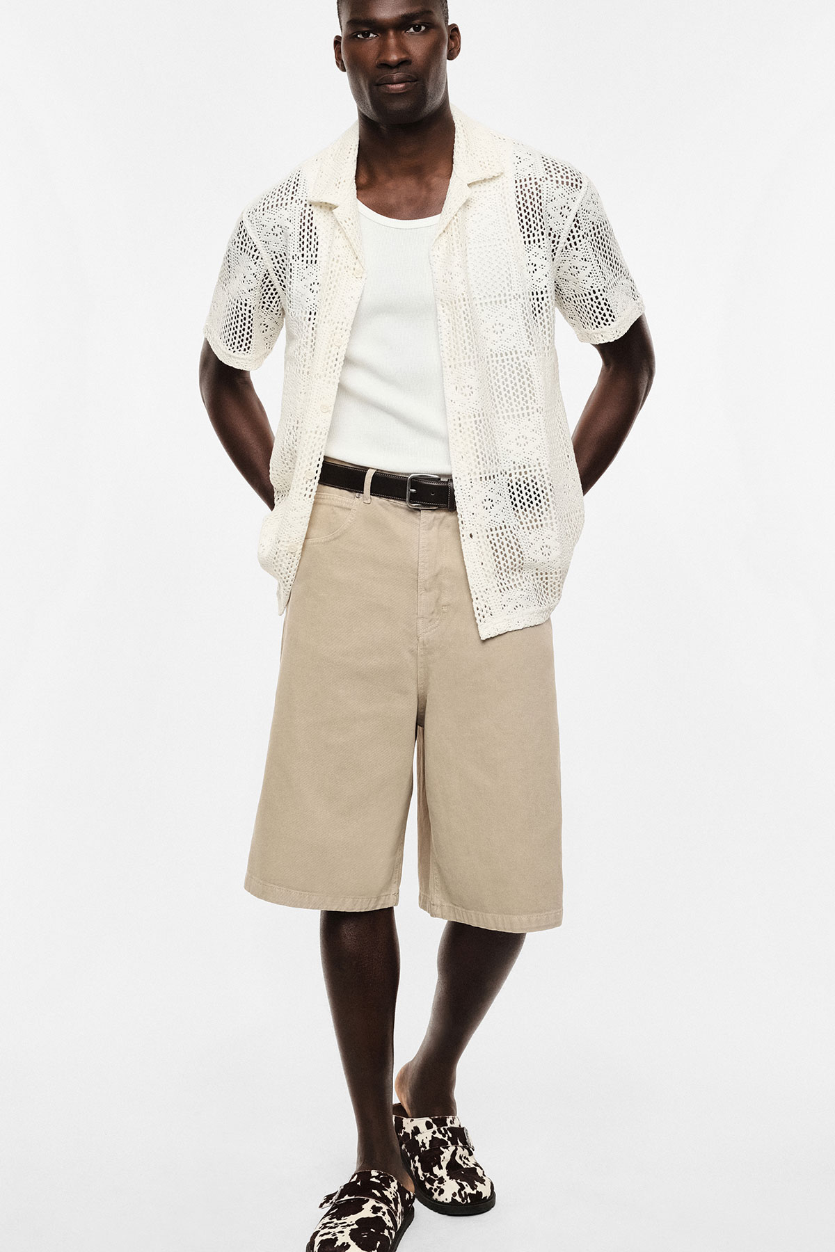 Model on white background wearing cream crochet shirt over white tank, beige long shorts, black belt and patterned slip-ons