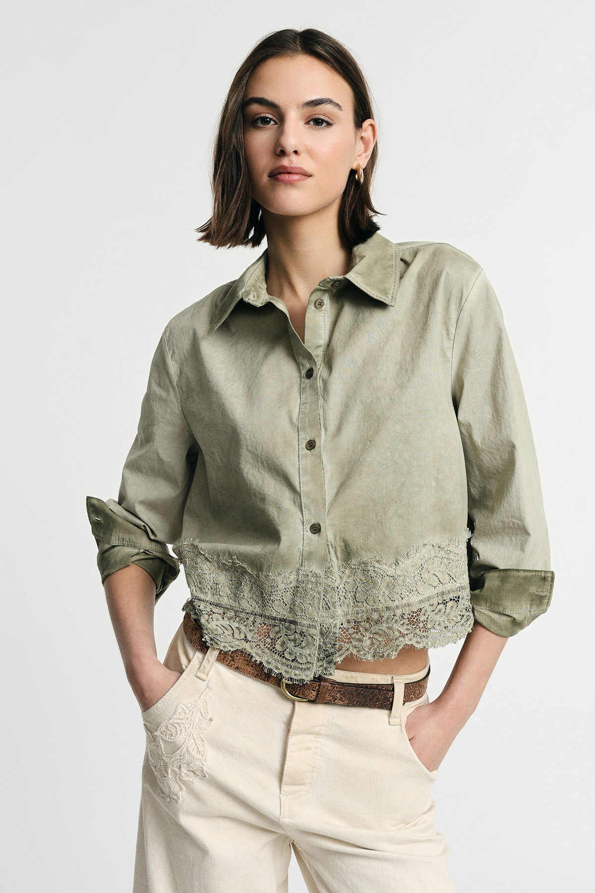 Woman wearing sage green button-up cropped shirt with lace hem and cream embroidered trousers, hands in pockets