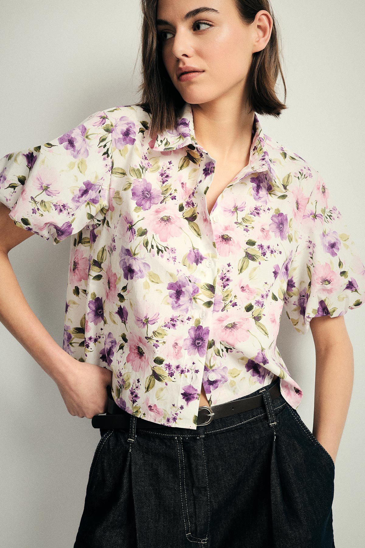 Woman wearing a white floral short-sleeve blouse with purple and pink flowers tucked into high-waist dark jeans, hands in pockets