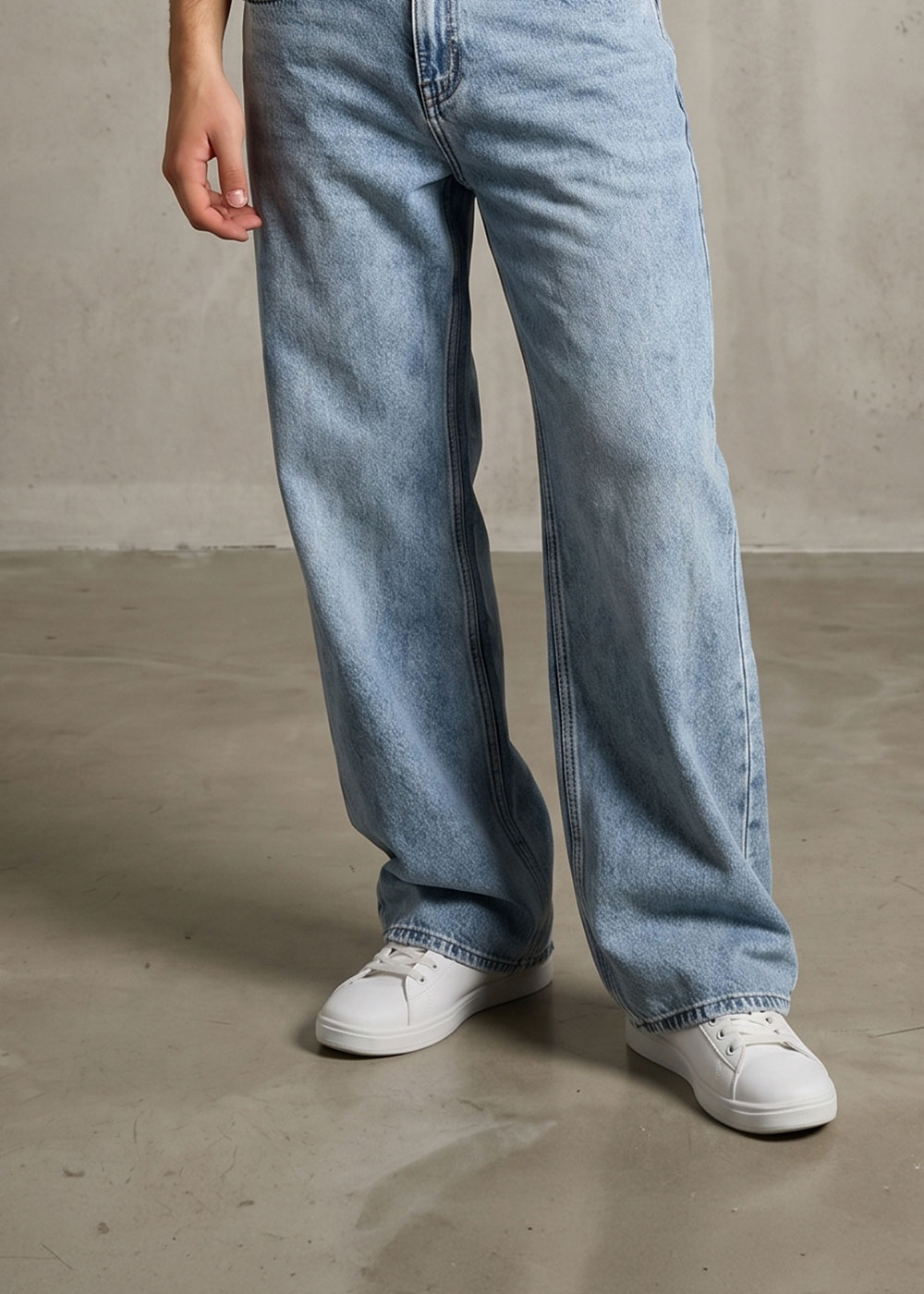 Lower body wearing light blue wide-leg jeans and white trainers on a concrete floor, casual styling with hand at side