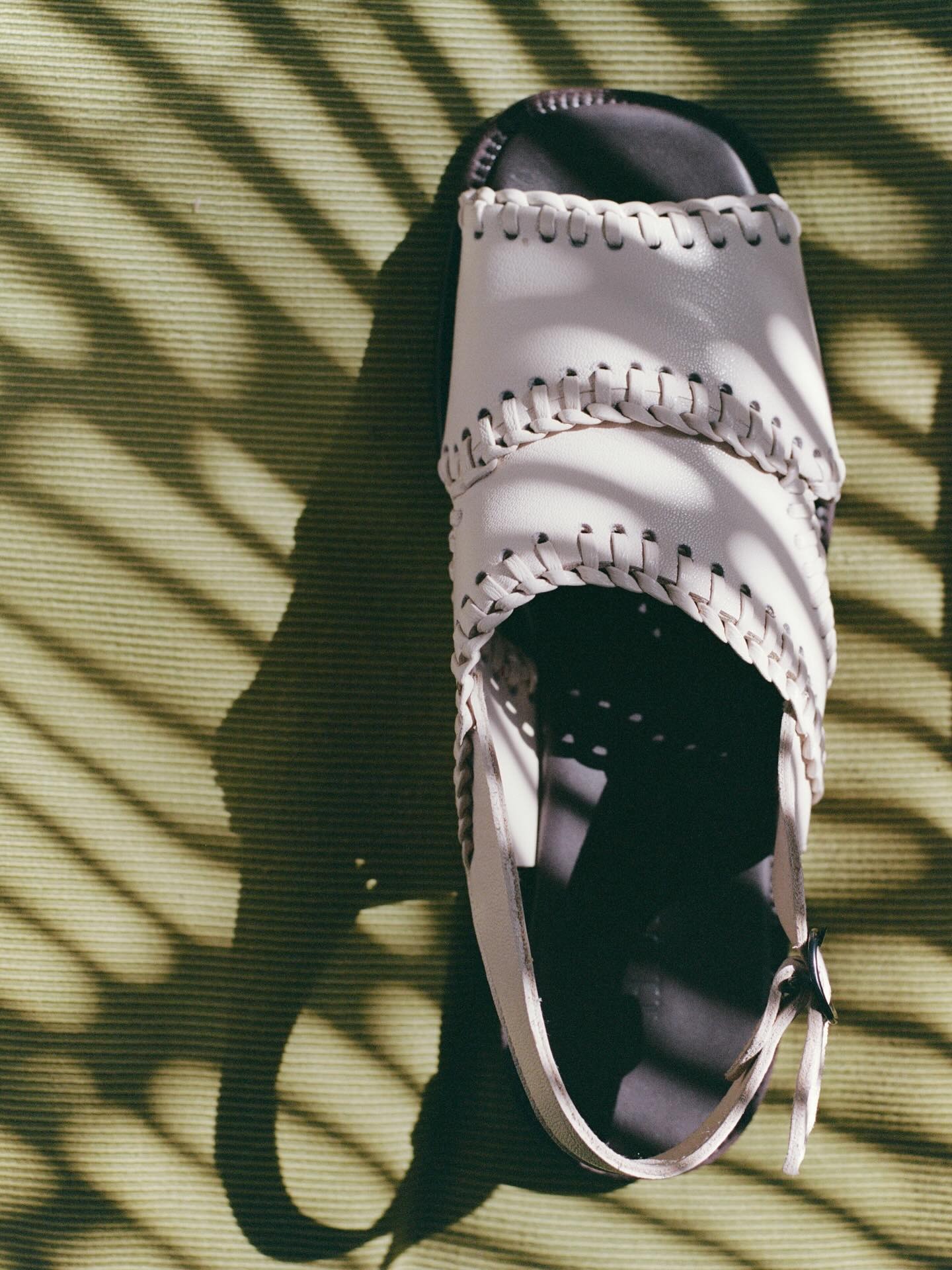 White leather woven-strap sandal with ankle buckle on green textured mat, striped shadow pattern