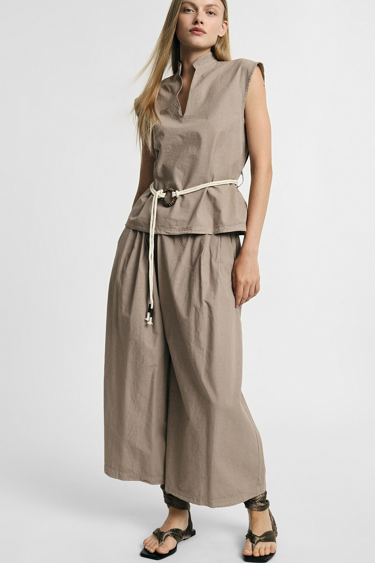 Woman wearing beige sleeveless top and wide-leg trousers with a rope belt and sandals on white background