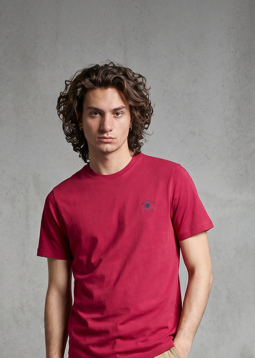 Young man with shoulder-length curly hair wearing a red crew-neck T-shirt, standing against a grey concrete backdrop with one hand in his pocket