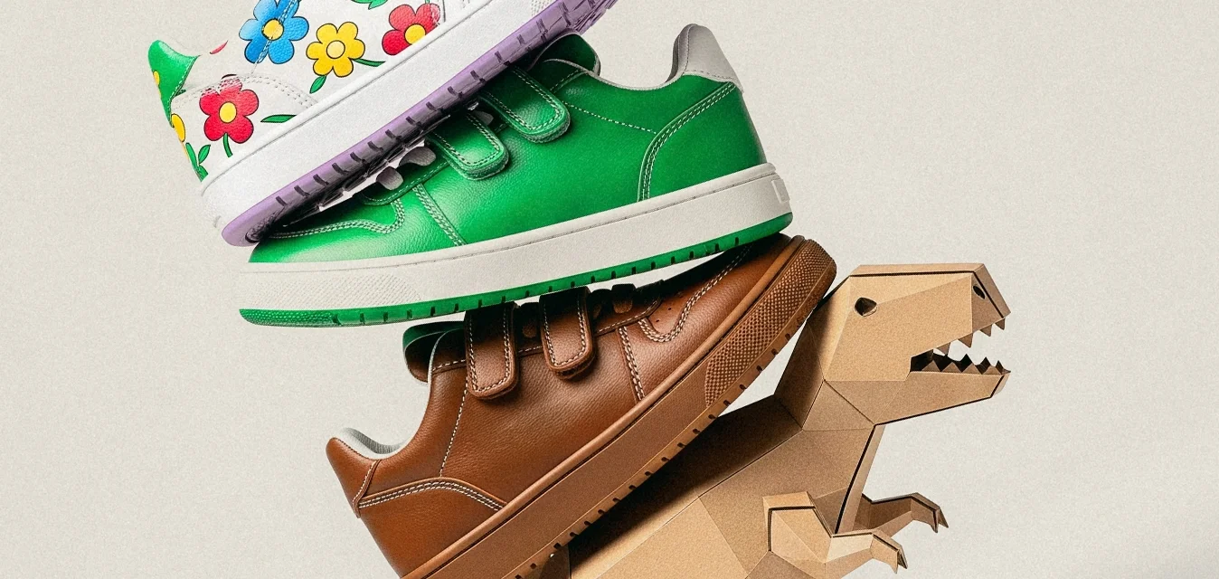 Scarpe di dimensioni infantili, bianche, verdi e marroni, impilate su una piccola figura di cartone a forma di dinosauro marrone, su uno sfondo beige semplice.