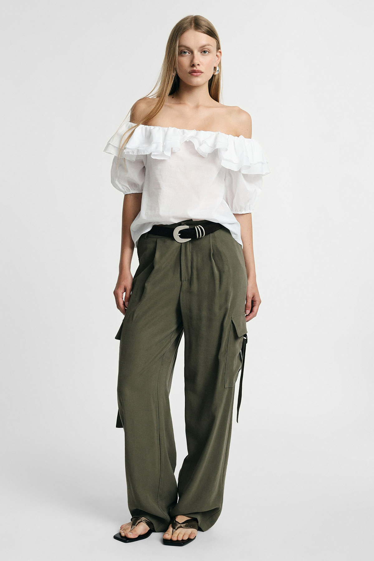 Female model in white off-the-shoulder ruffle blouse, olive cargo trousers with black belt and sandals, standing against plain background