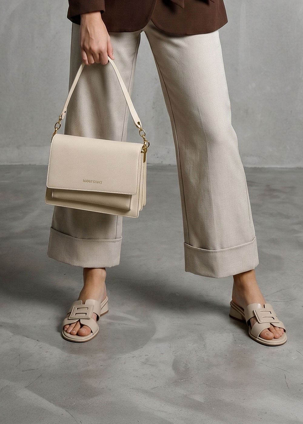 Woman wearing beige wide-leg trousers and slide sandals holding a cream leather shoulder bag on a concrete floor