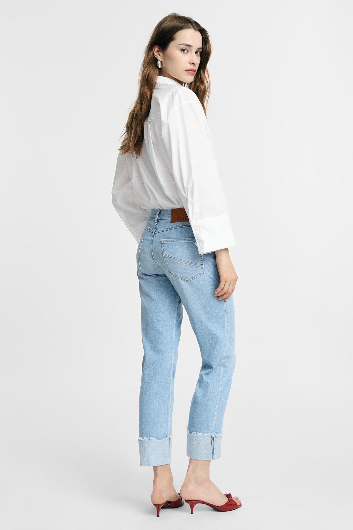 Woman in white shirt and cuffed light-blue jeans wearing red heeled mules, looking over her shoulder against a plain studio background