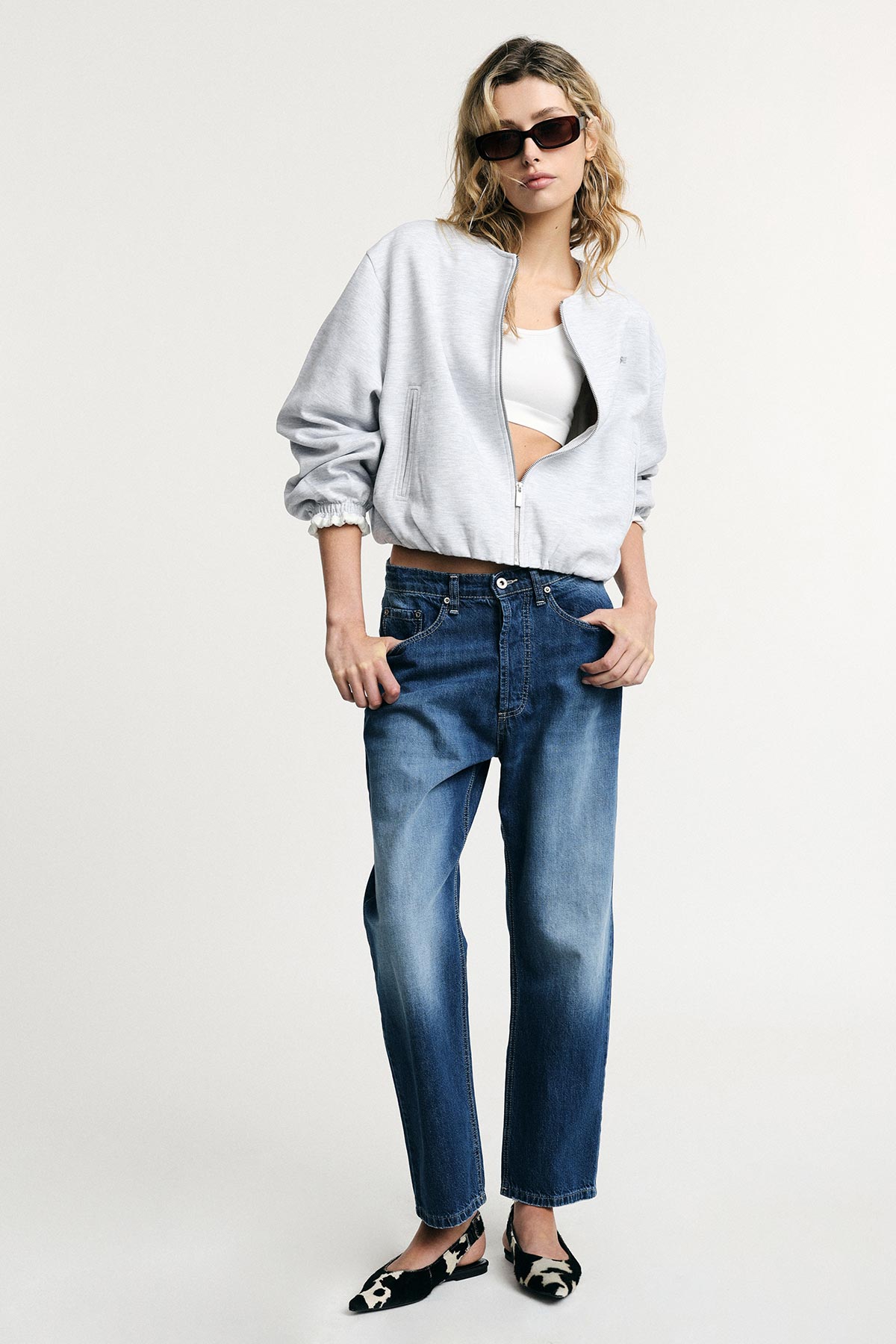 Model in grey zip bomber jacket, white crop top, relaxed blue jeans and patterned black flats, wearing sunglasses