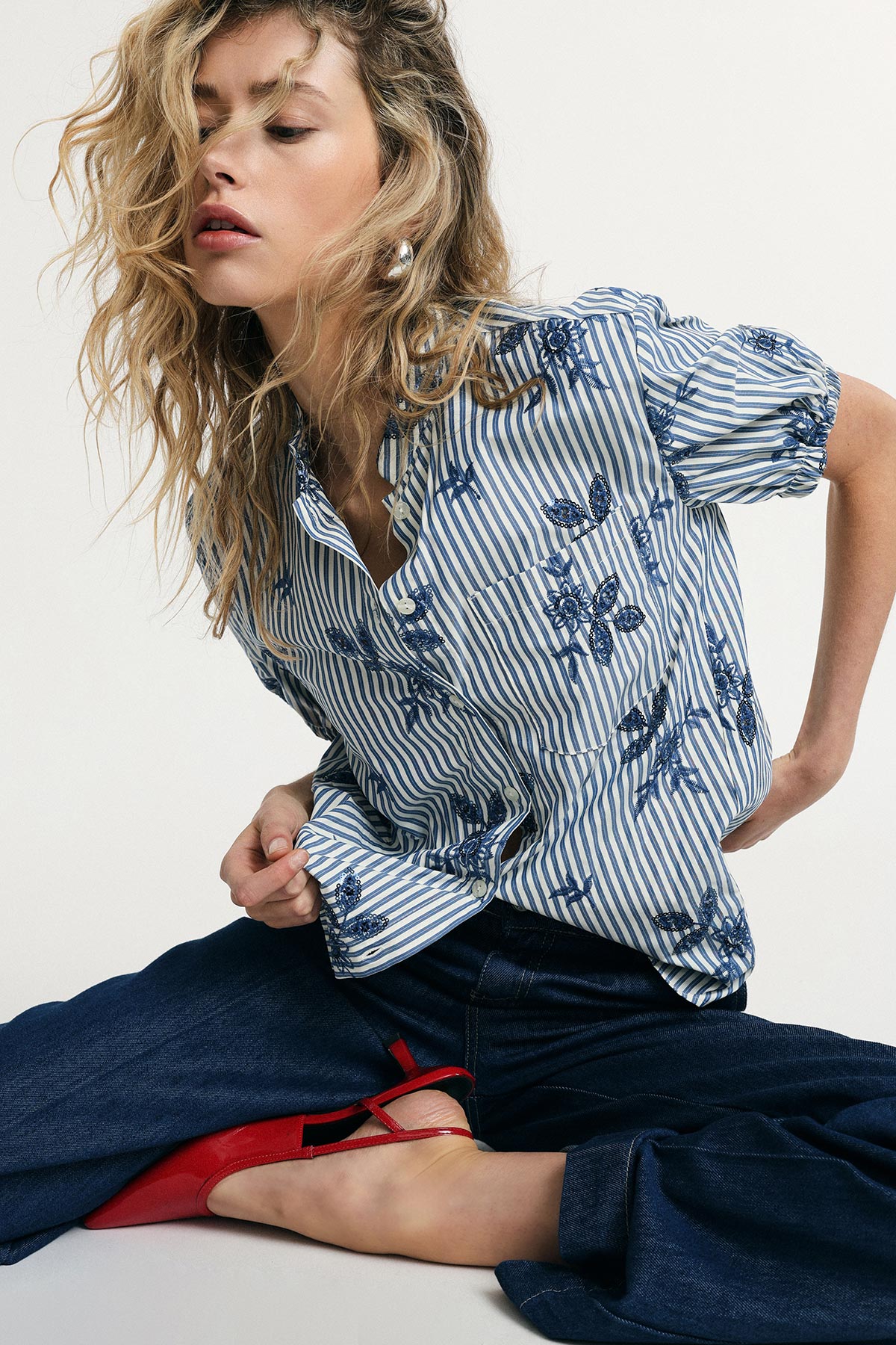 Woman modelling blue-and-white striped embroidered blouse, wide-leg jeans and red flats in seated fashion pose