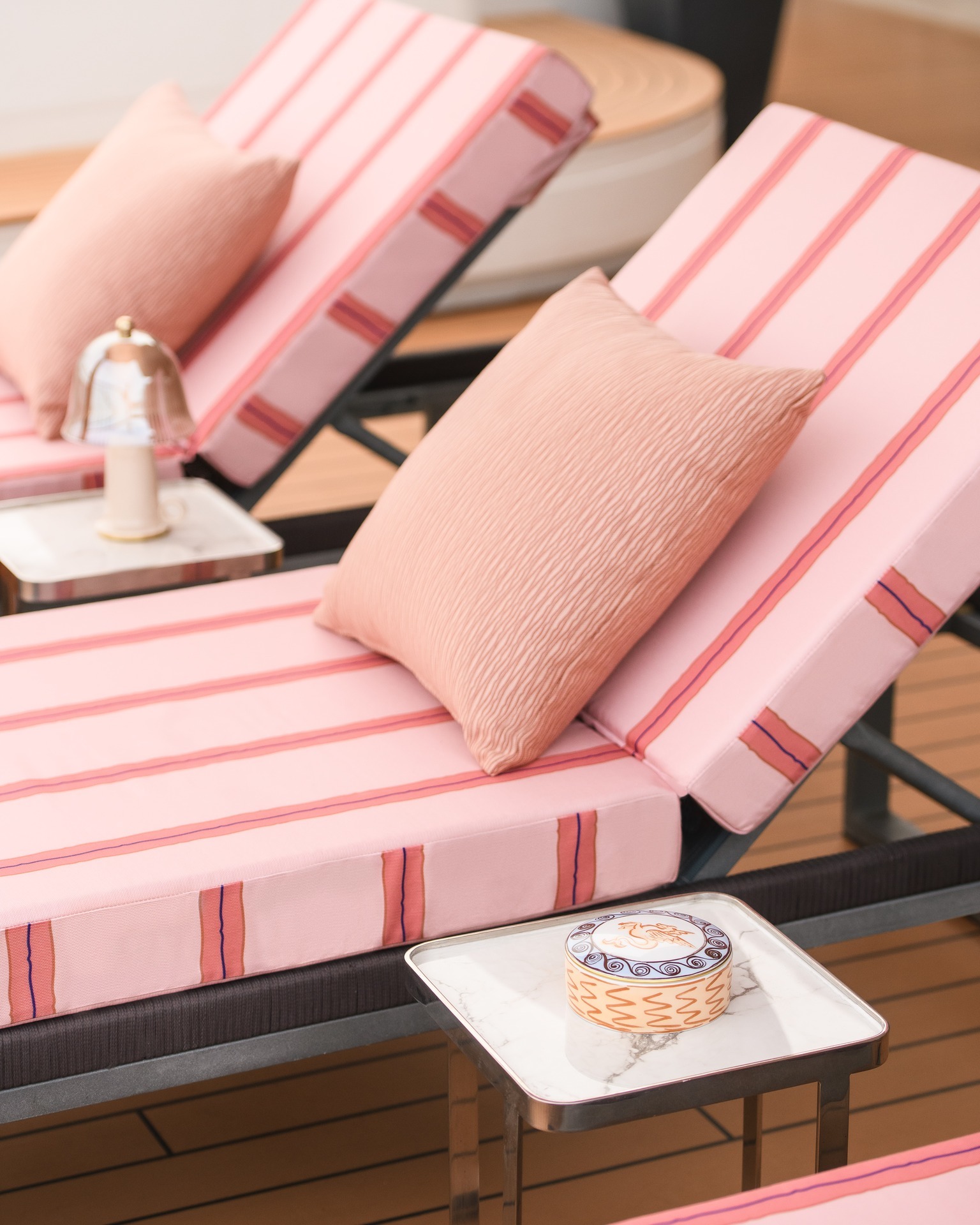 Two pink striped sunloungers with coral cushions on a wooden deck, marble side table with decorative tin