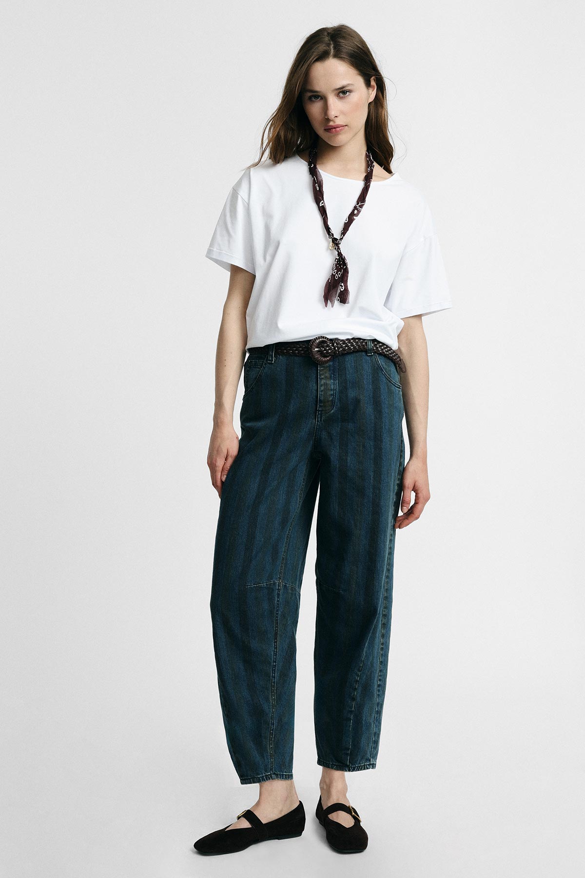 Woman modelling casual outfit: white oversized T-shirt, high-waisted relaxed blue jeans with braided belt, neck scarf and black flats