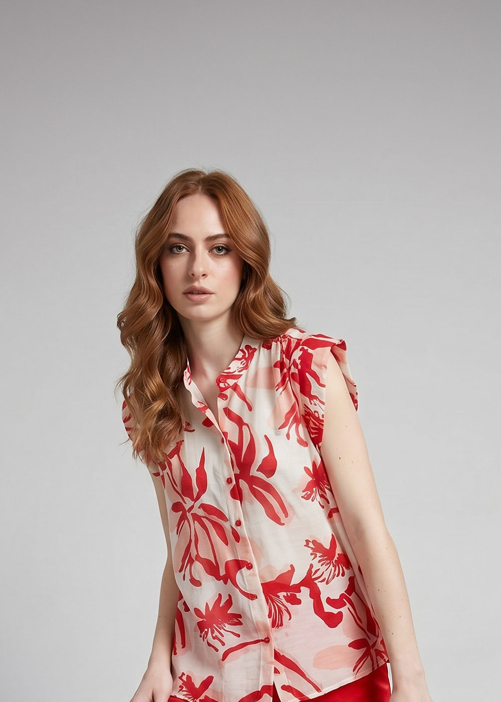 Red-haired woman wearing a sleeveless white blouse with bold red floral print, posing against a plain grey background