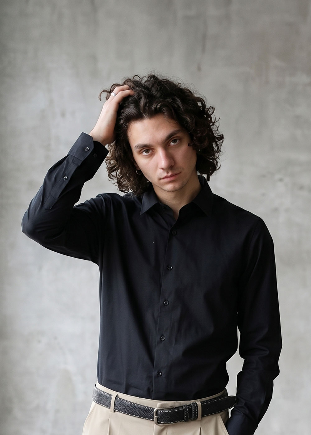 Portrait of a young man with curly hair in a black shirt and beige trousers, hand in hair against a grey backdrop