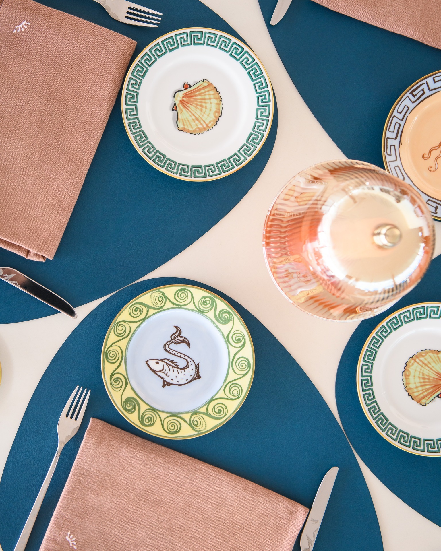 Top-down view of elegant table settings with decorative shell and fish plates, teal placemats, pink napkins, cutlery and glass