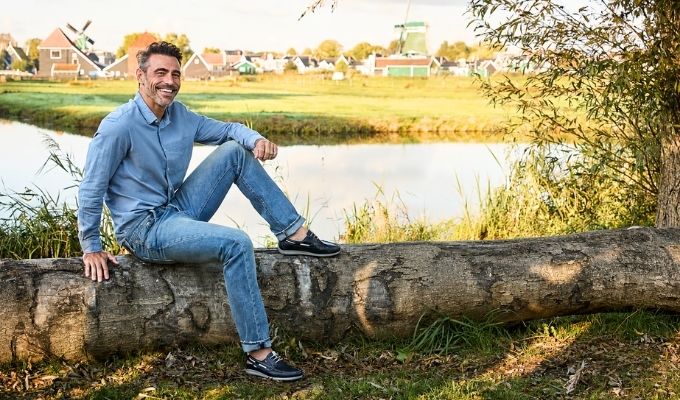 Smiling man sitting on a log beside a canal with Dutch windmills and a village in the background