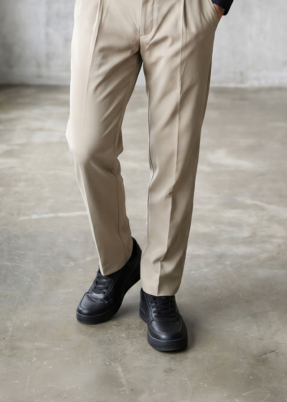 Lower body of person wearing tailored beige trousers and black trainers, standing on a polished concrete floor