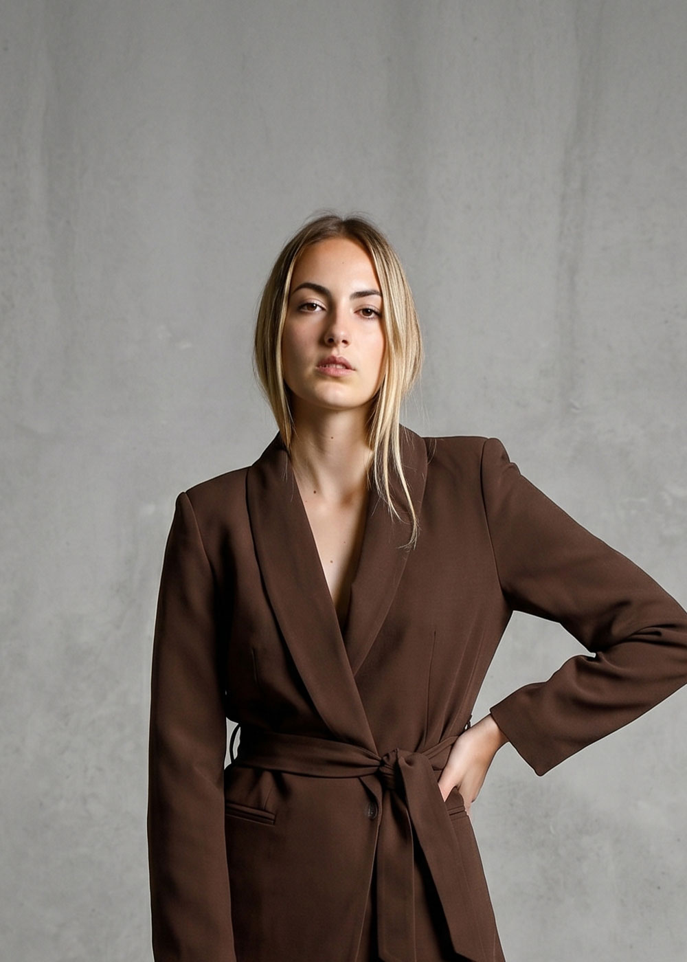 Young woman wearing a brown belted blazer posing against a grey concrete wall