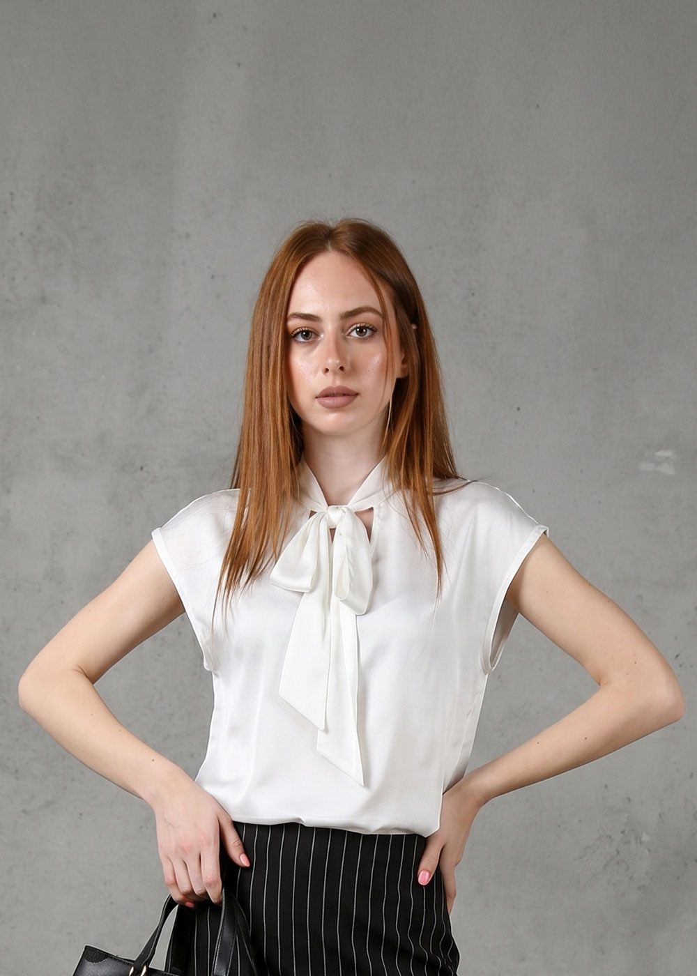 Young woman model in white bow-neck blouse and black pinstripe skirt holding a small black bag against a grey concrete wall