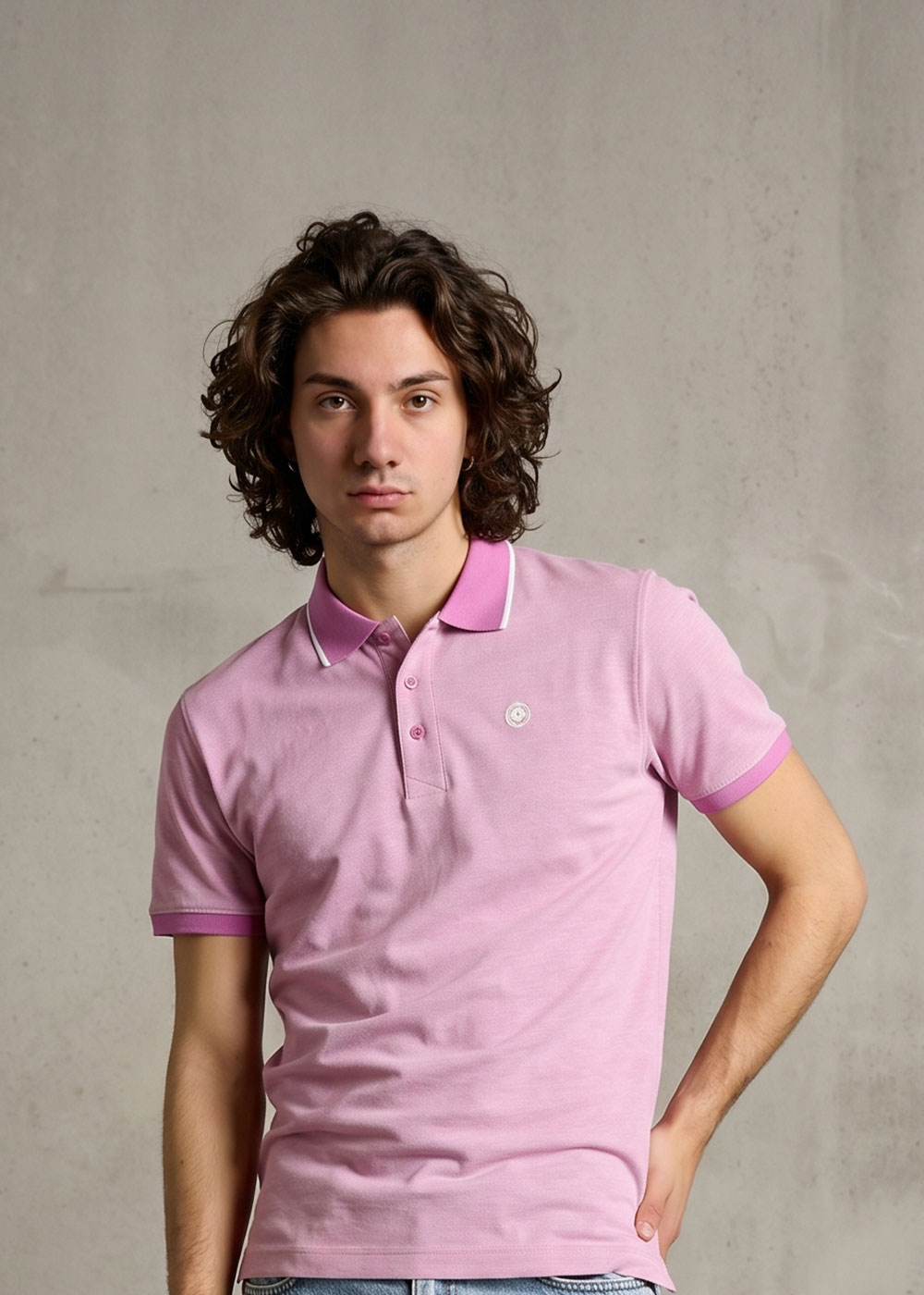 Young man with curly hair wearing a pink polo shirt standing against a concrete wall