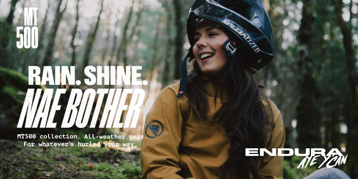 Smiling cyclist in helmet and yellow waterproof MT500 jacket in woodland, promotional text 'Rain. Shine. NAE BOTHER' with Endura logo