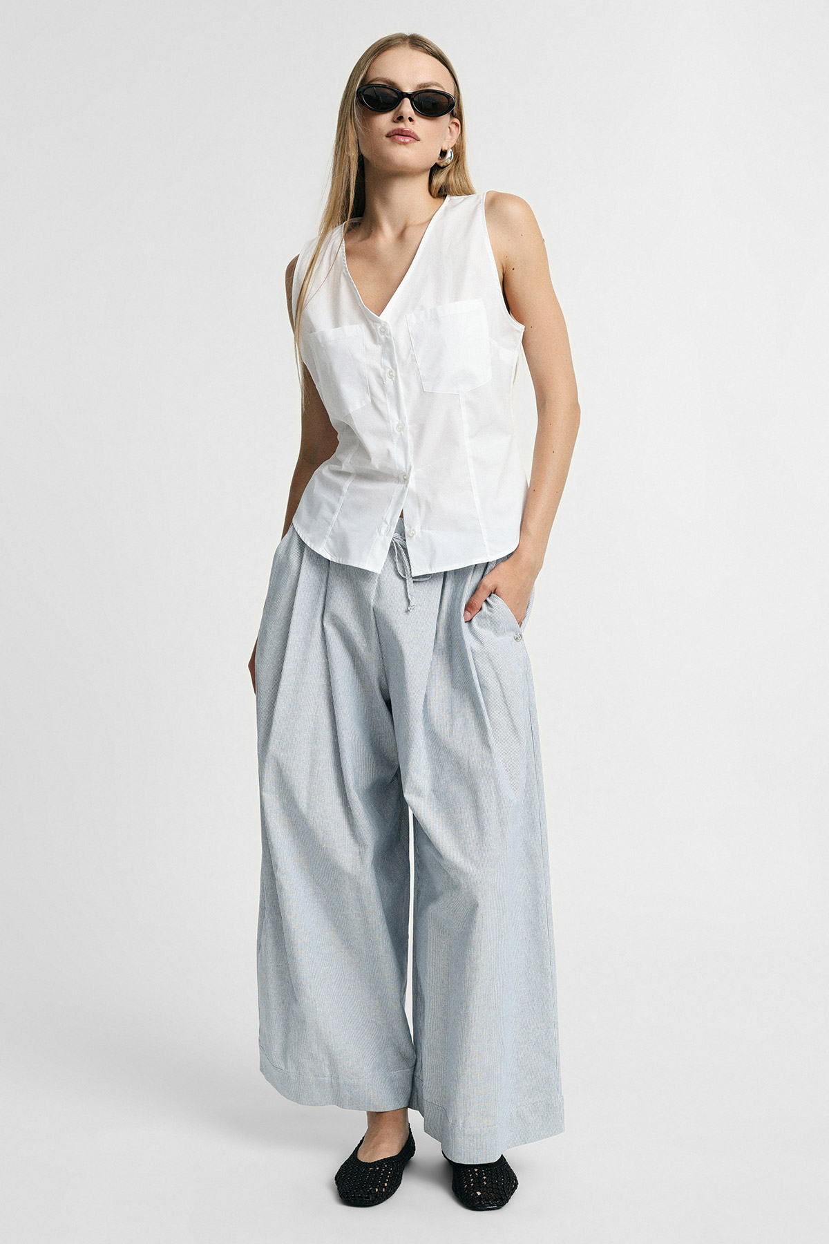 Female model in white sleeveless button-front blouse and wide-leg pale grey striped trousers, black flats and sunglasses