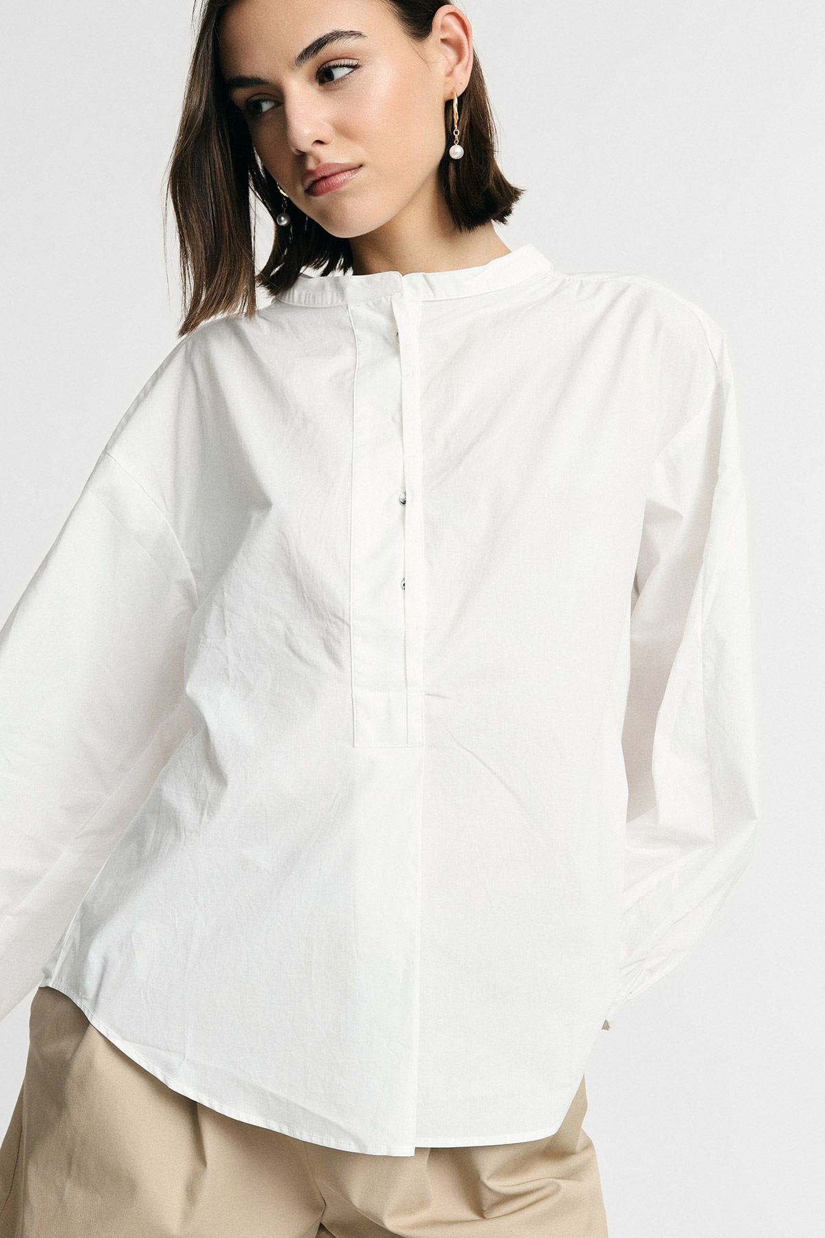 Woman wearing white long-sleeve buttoned mandarin-collar blouse and beige trousers, pearl drop earrings, studio portrait