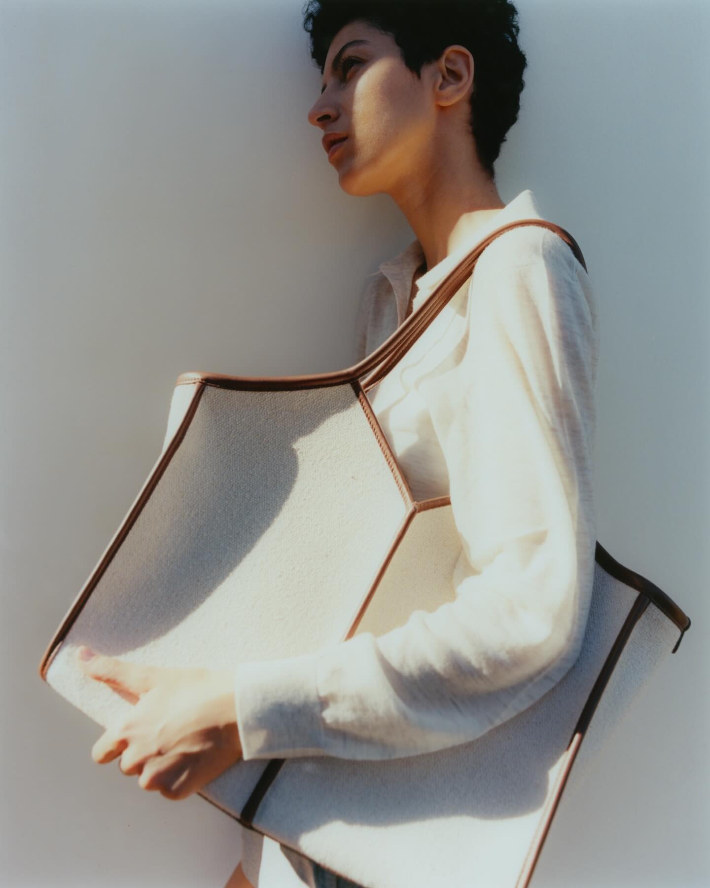 Person in white shirt, side-profile portrait holding large cream canvas tote bag with brown trim against a pale background