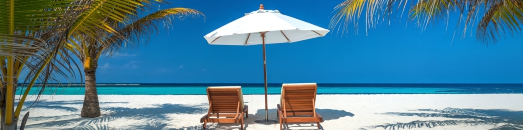 Two wooden sun loungers under a white parasol on a white-sand tropical beach with turquoise sea and palm trees