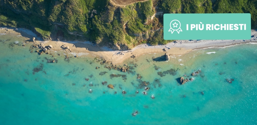 Aerial view of turquoise sea meeting a sandy beach and rocky shoreline beneath green cliffs, teal badge reading "I PIÙ RICHIESTI" top-right