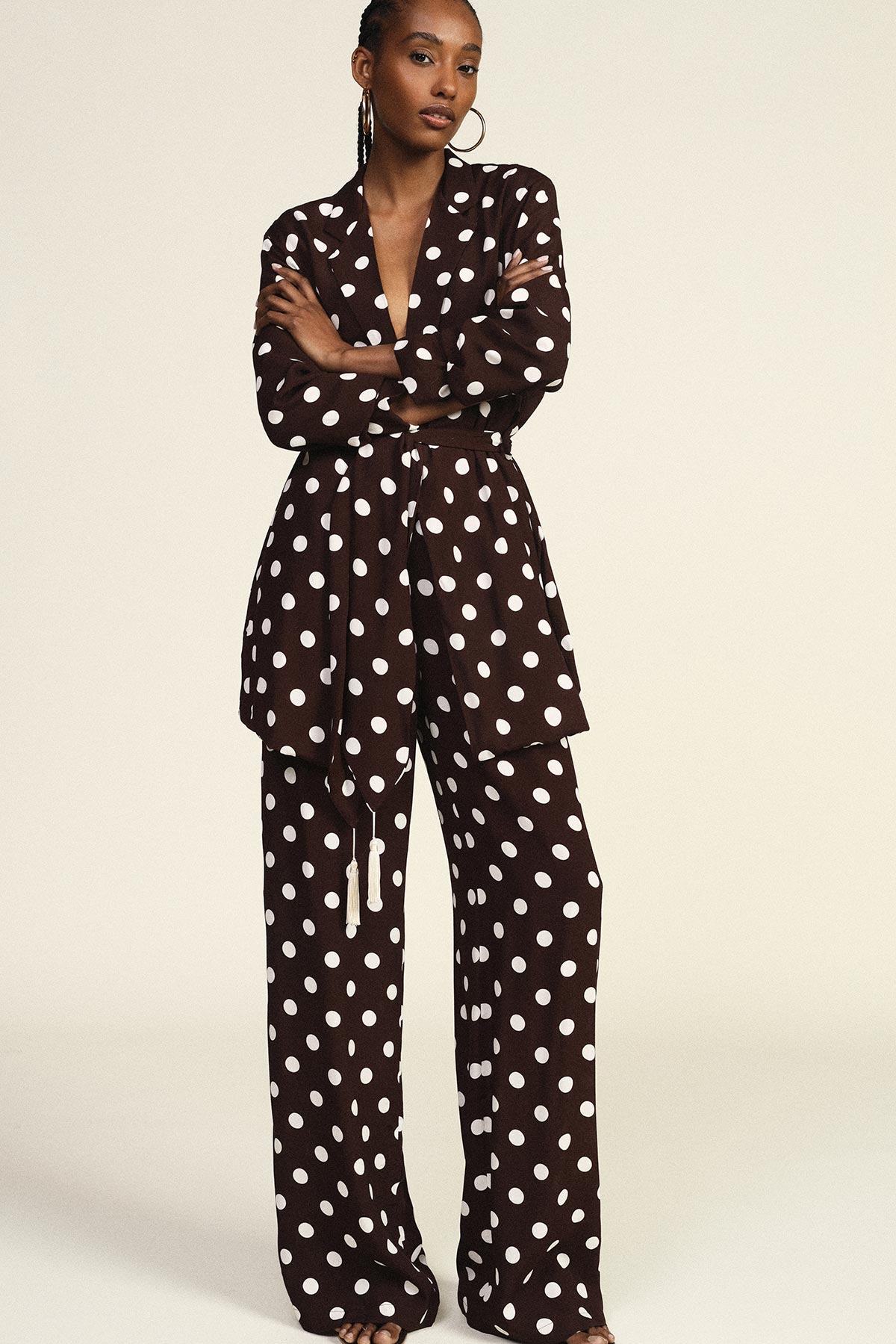 Model wearing brown suit with large white polka dots: belted blazer, wide-leg trousers and gold hoop earrings on neutral background