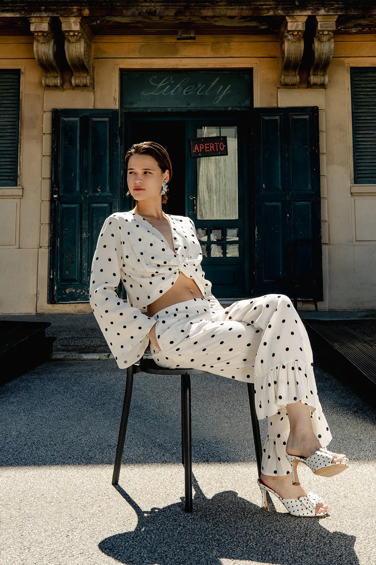 Woman in white polka-dot co-ord and heels seated on a chair in front of a vintage dark green doorway with a 'Liberty' sign