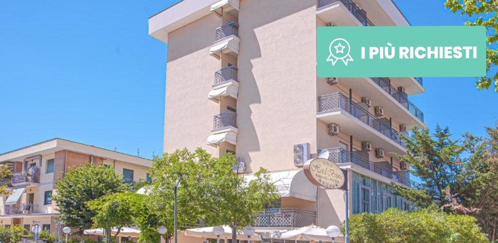Exterior of a pale-coloured seaside hotel with multiple balconies, outdoor umbrellas and trees beneath a clear blue summer sky