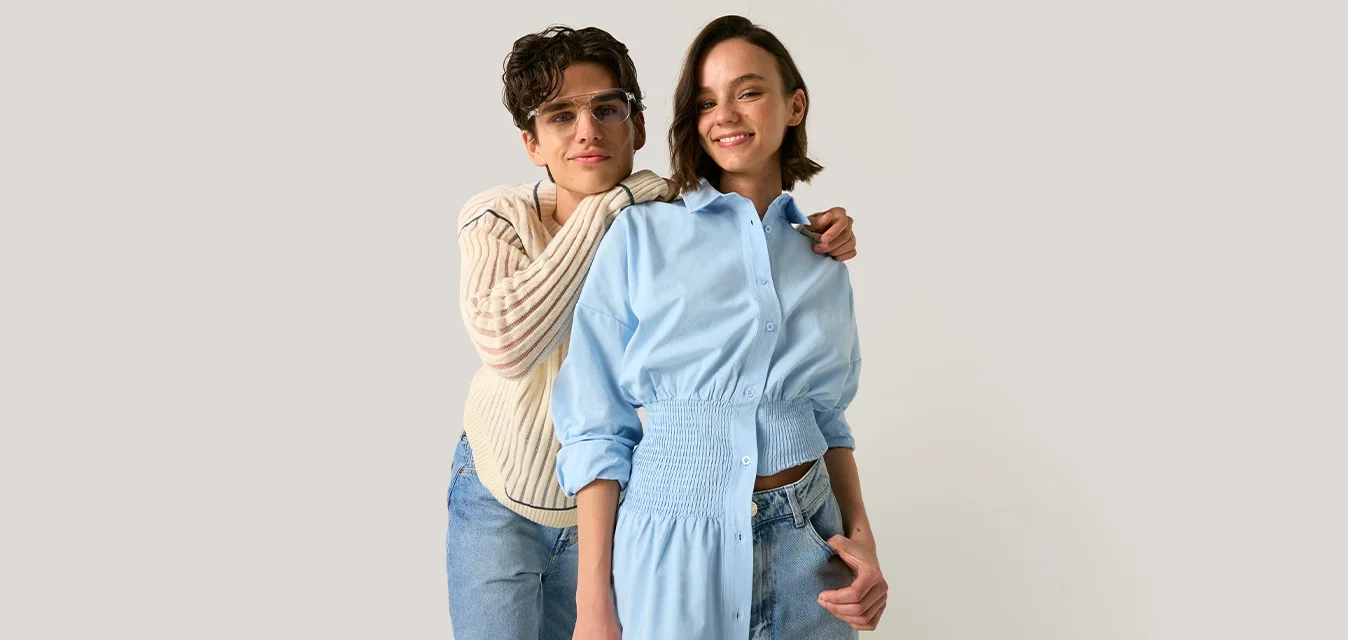 Due giovani donne stanno vicine, una con occhiali e maglione crema abbraccia un'altra con un vestito camicia blu, entrambe sorridenti su uno sfondo semplice.