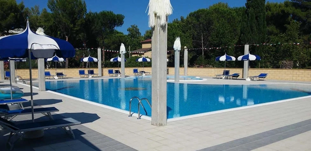 Outdoor rectangular swimming pool with blue sun umbrellas and loungers on a sunlit tiled terrace, surrounded by trees and clear sky