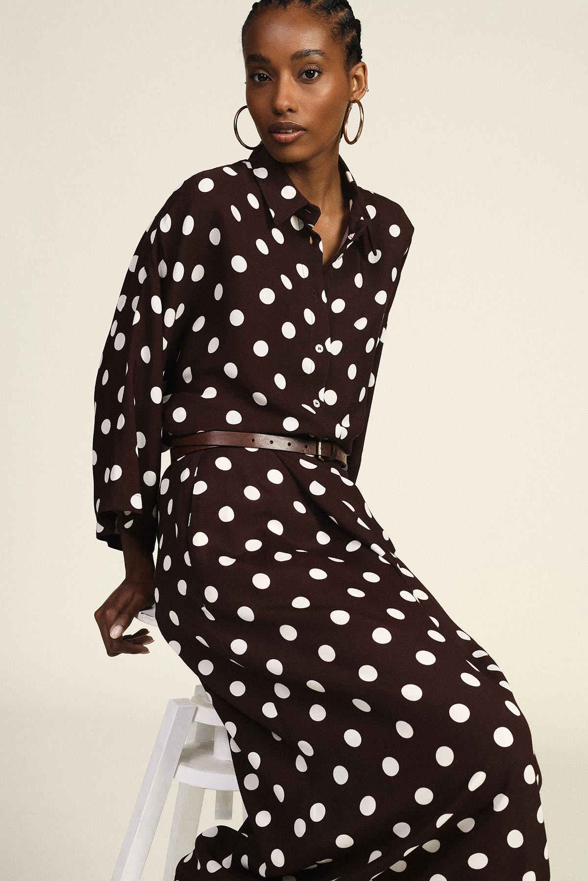 Model in brown polka-dot dress with belt and hoop earrings, seated on white stool against pale background