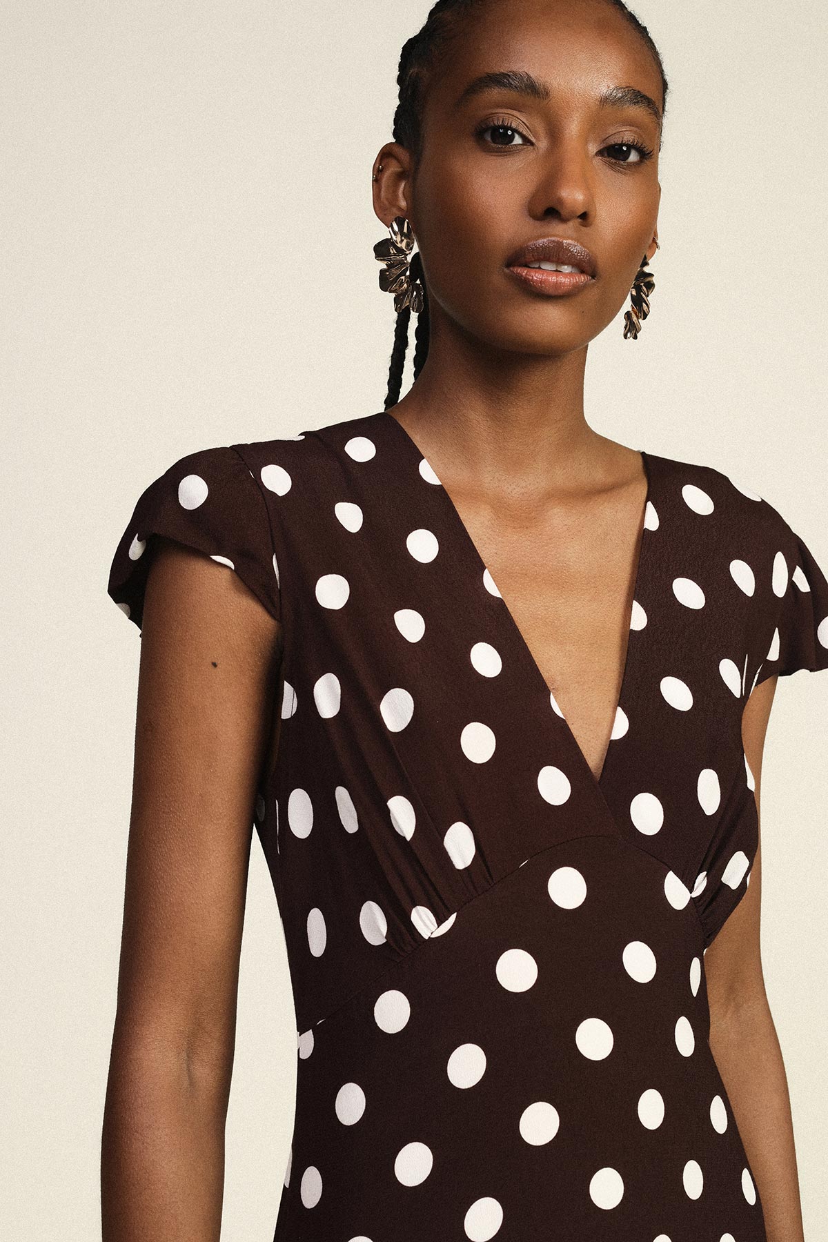 Woman wearing brown V-neck polka dot dress and gold statement earrings, posing against a neutral background
