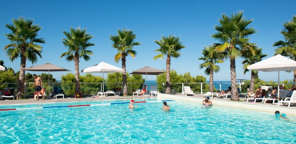 Outdoor swimming pool with families and sunloungers under umbrellas, palm trees and sea view on a sunny day