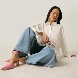 Donna seduta sul pavimento che indossa una camicetta bianca con bordo in pizzo, jeans a gamba larga e scarpe con tacco rosa, su uno sfondo chiaro e semplice.