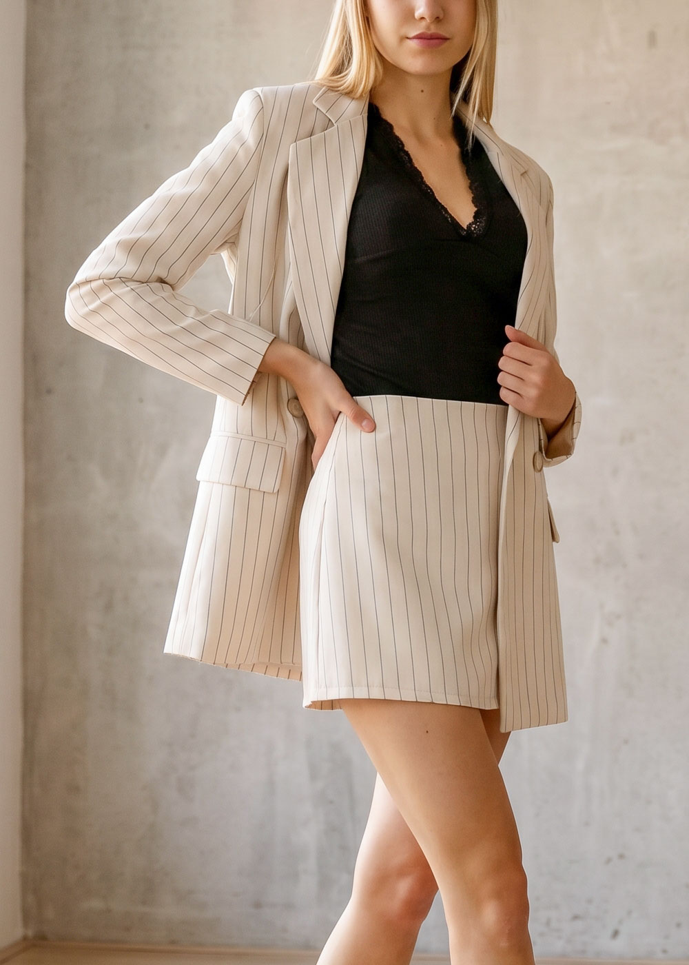 Model in cream pinstripe blazer and matching shorts with black lace-trim camisole, standing against a neutral concrete wall