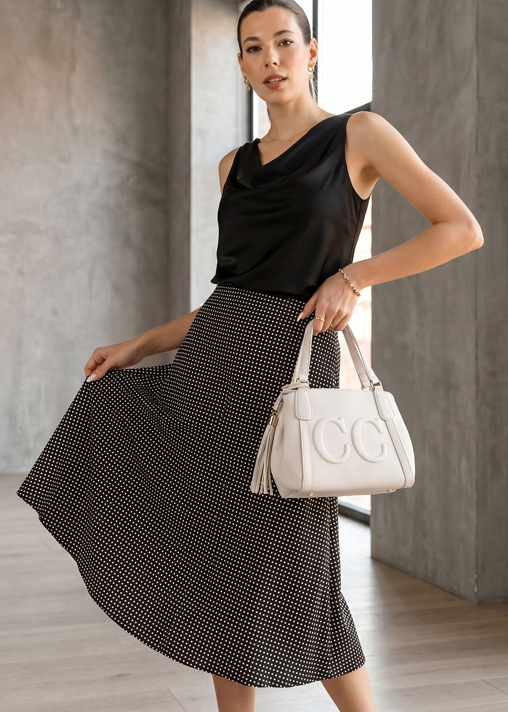 Woman in black satin sleeveless top and polka-dot midi skirt holding a white embossed handbag in a minimalist interior