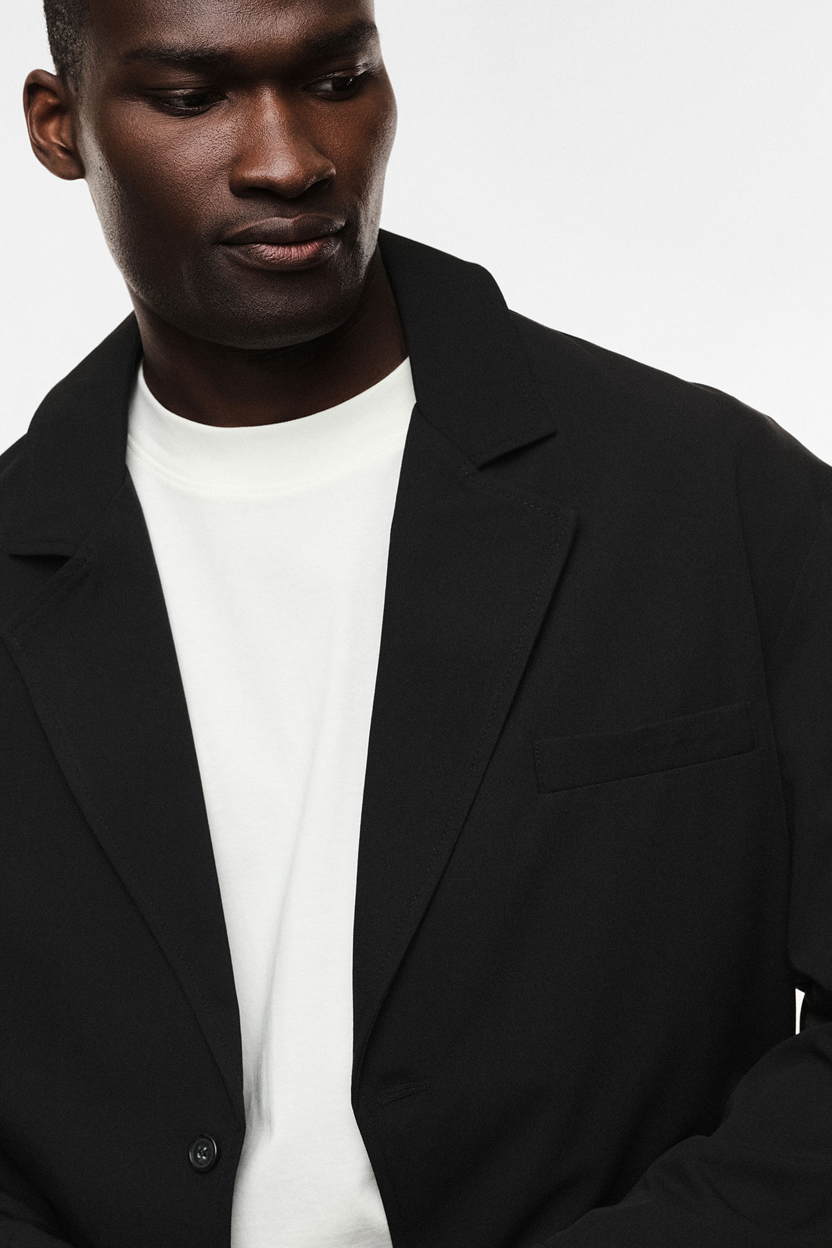 Close-up studio portrait of a person wearing a black blazer over a white t-shirt, looking down, men's fashion