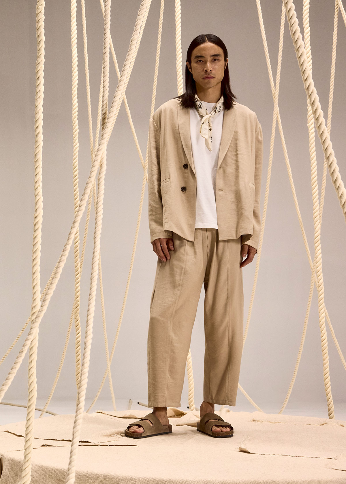 Model in beige relaxed suit, white tee and scarf with sandals, standing amidst hanging thick ropes on a neutral studio backdrop
