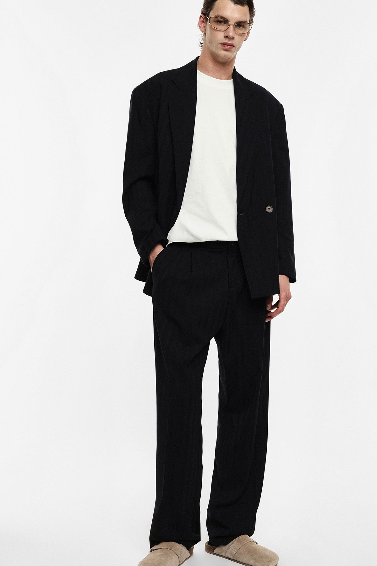 Male model wearing oversized black suit and white T-shirt, beige suede shoes and clear glasses, standing against white background