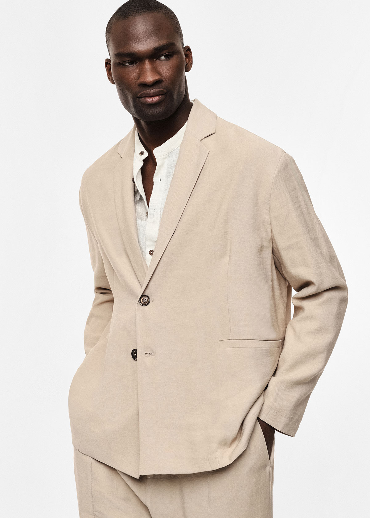 Male model wearing a beige suit and white linen shirt, hands in pockets, looking to the side against a plain white background