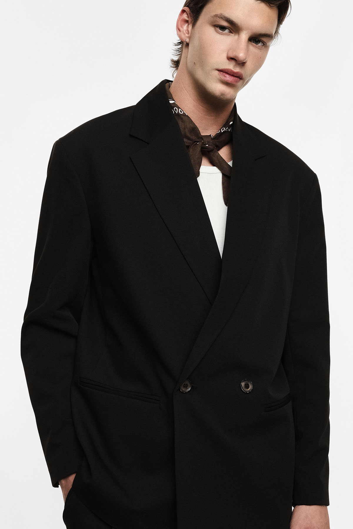 Male fashion portrait: model wearing a black double-breasted blazer, white ribbed top and brown neck scarf on white background