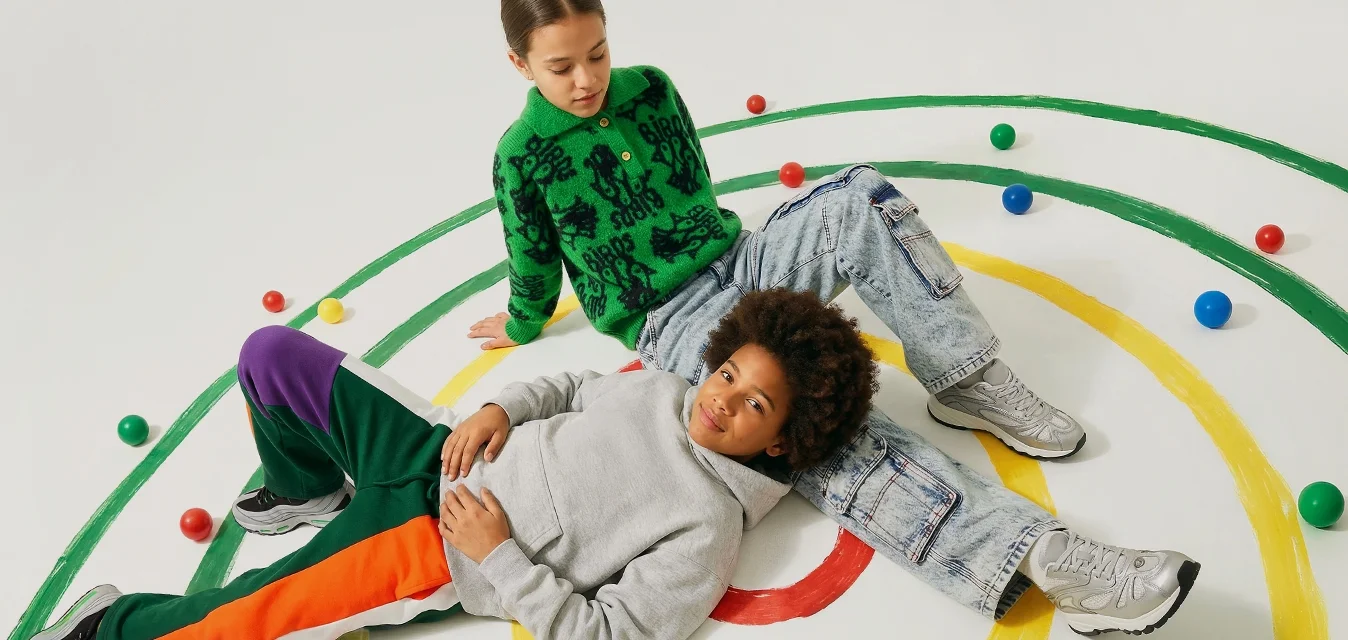 Due bambini su un pavimento bianco con cerchi concentrici gialli e verdi, circondati da palline colorate sparse.