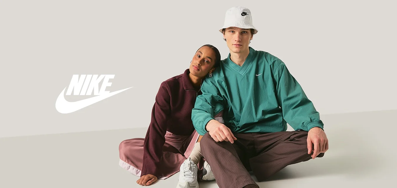 Due giovani adulti seduti sul pavimento, uno appoggia la testa sulla spalla dell'altro, entrambi indossano abbigliamento sportivo casual e sneakers.
