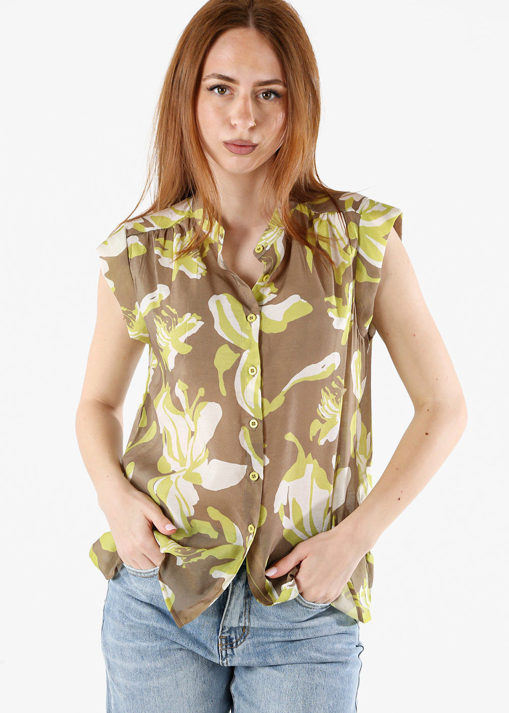 Woman wearing sleeveless brown and lime floral button-up blouse and blue jeans, hands in pockets, neutral expression